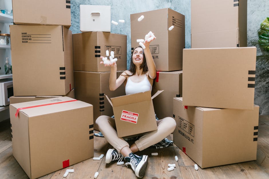 A person’s hands are shown packing a large brown cardboard box, writing the word 'KITCHEN' in black marker on the side with a small heart symbol. The individual is wearing a wristwatch and holding t